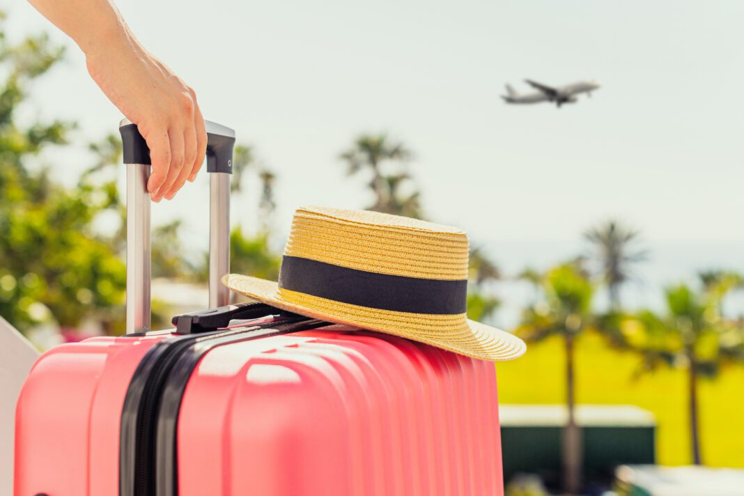 Woman with pink suitcase and beach hat standing on passengers ladder and getting out of airplane opposite sea coastline with palm trees. Tourism concept